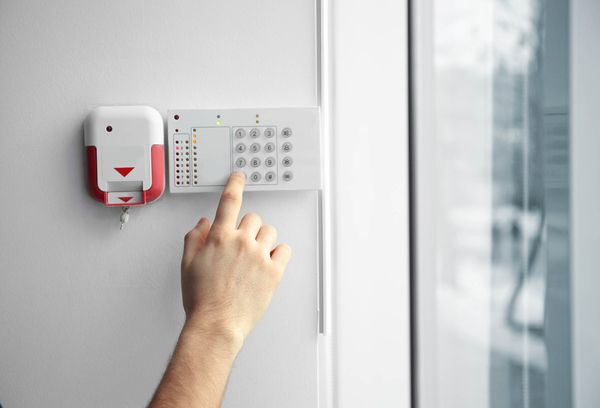Eine Hand drückt eine Taste auf einem weißen Sicherheitssystem mit einem Ziffernfeld und einer roten Alarmanlage an der Wand.
