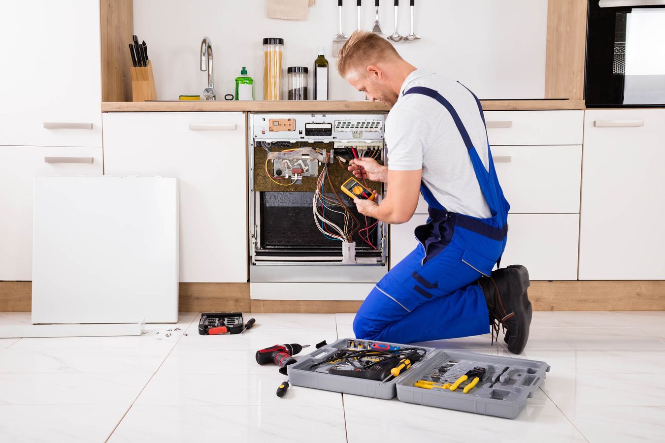 Ein Techniker in blauer Arbeitskleidung kniet vor einem geöffneten Elektrogerät in einer modernen Küche. Er hält ein Messgerät in der Hand und arbeitet an den Kabeln im Inneren des Geräts. Neben ihm liegt ein Werkzeugkasten mit verschiedenen Werkzeugen.