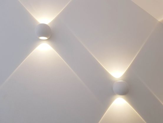 Zwei runde, weiße Wandleuchten, die warmes Licht abstrahlen und diagonale Lichtstrahlen auf die Wand werfen.