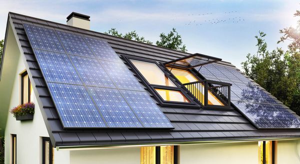 Dach eines Hauses mit mehreren Solarpanelen, darunter ein Fenster mit offener Klappe. Umgeben von Bäumen und einem klaren Himmel.