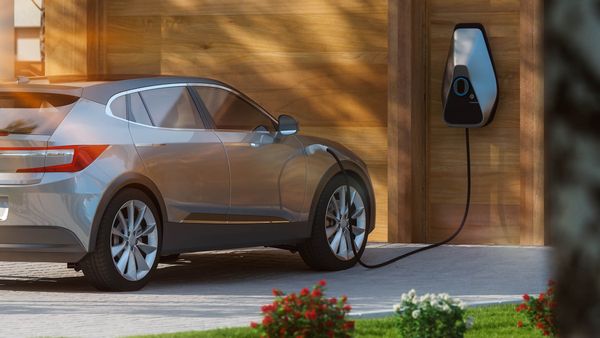 Ein silbernes Elektroauto parkt neben einer Holzgarage und ist an eine Ladestation angeschlossen. Die Ladestation hat eine ovale Form mit einem blauen Licht.