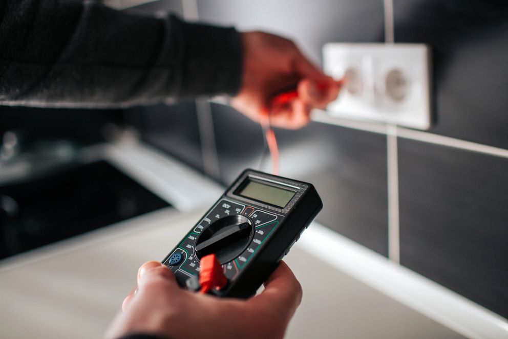 Eine Person hält ein Multimeter in der Hand und misst eine Steckdose. Das Multimeter hat einen digitalen Bildschirm und rote sowie schwarze Messleitungen.