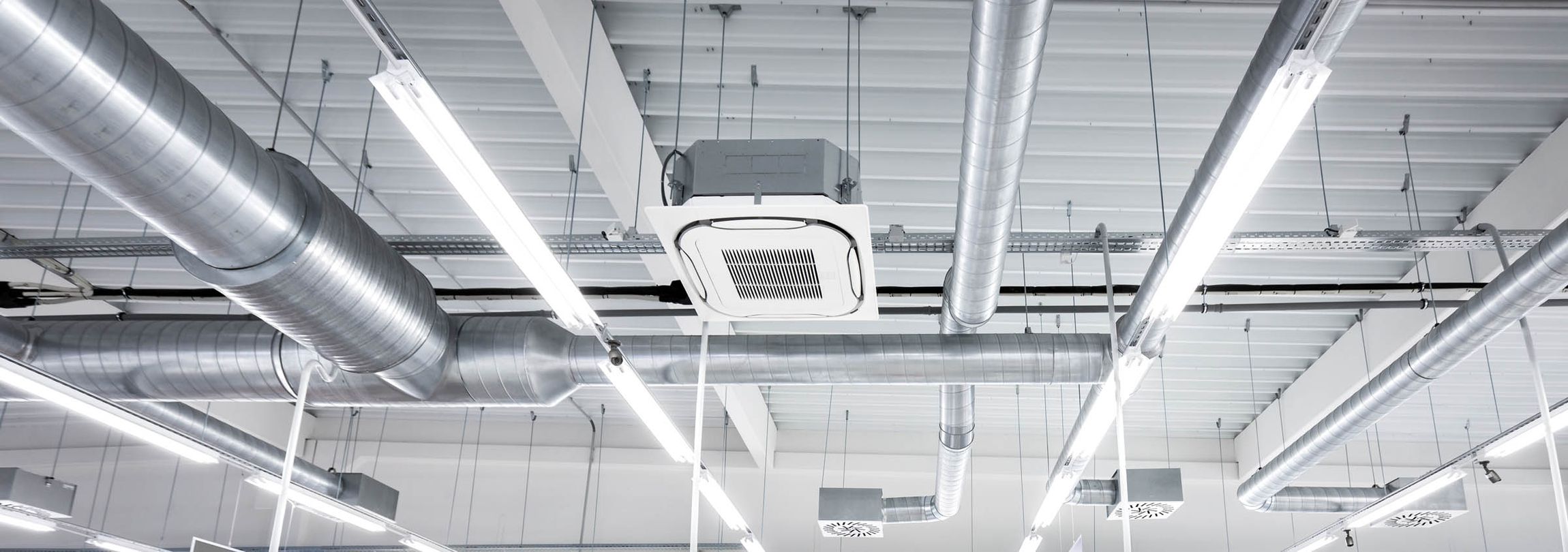 Decke eines Raumes mit mehreren silbernen Lüftungsrohren und einer Deckenklimaanlage in der Mitte. Helle LED-Leuchten sind entlang der Decke angebracht.